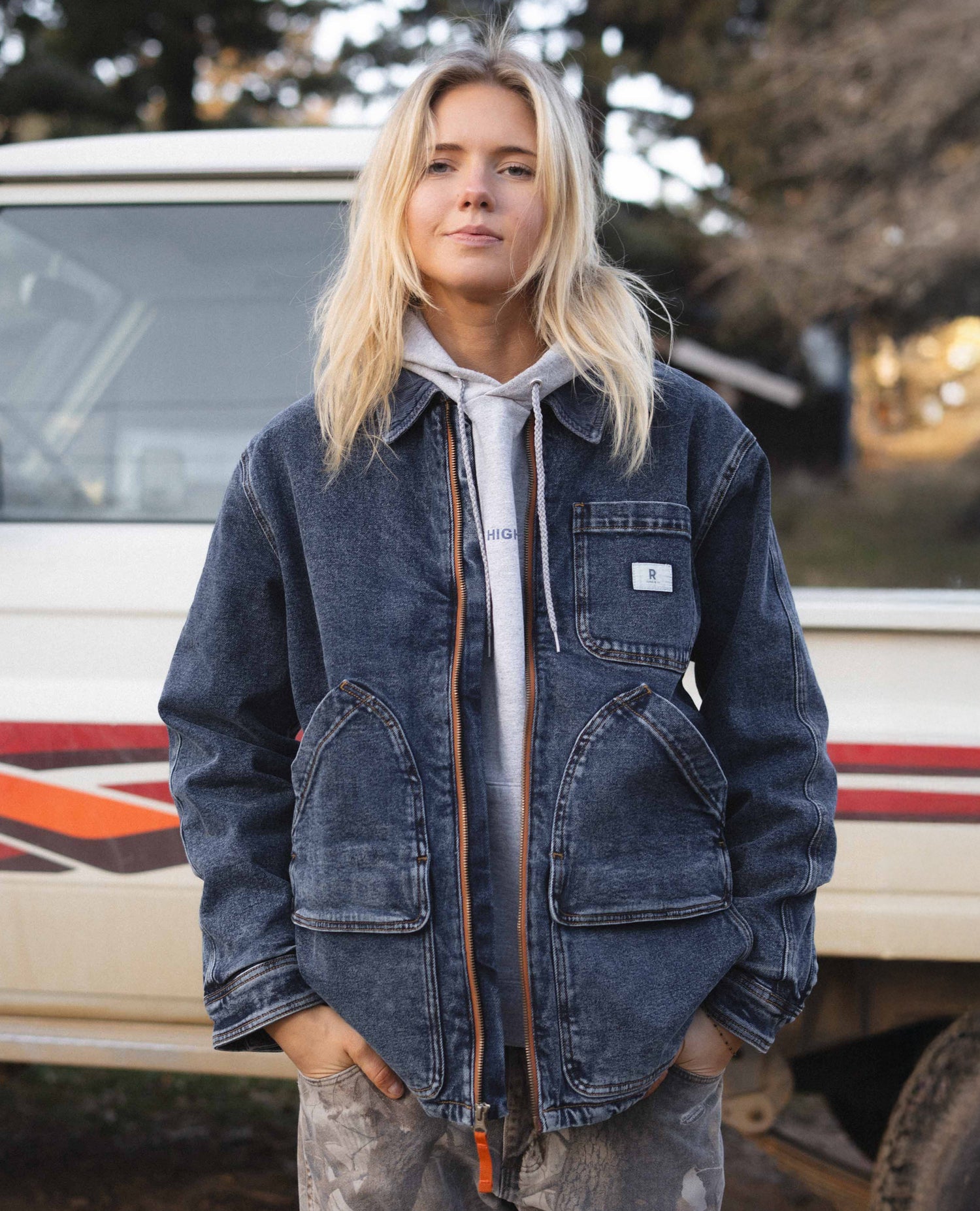 Person wearing a blue denim jacket standing in front of a vehicle with trees in the background