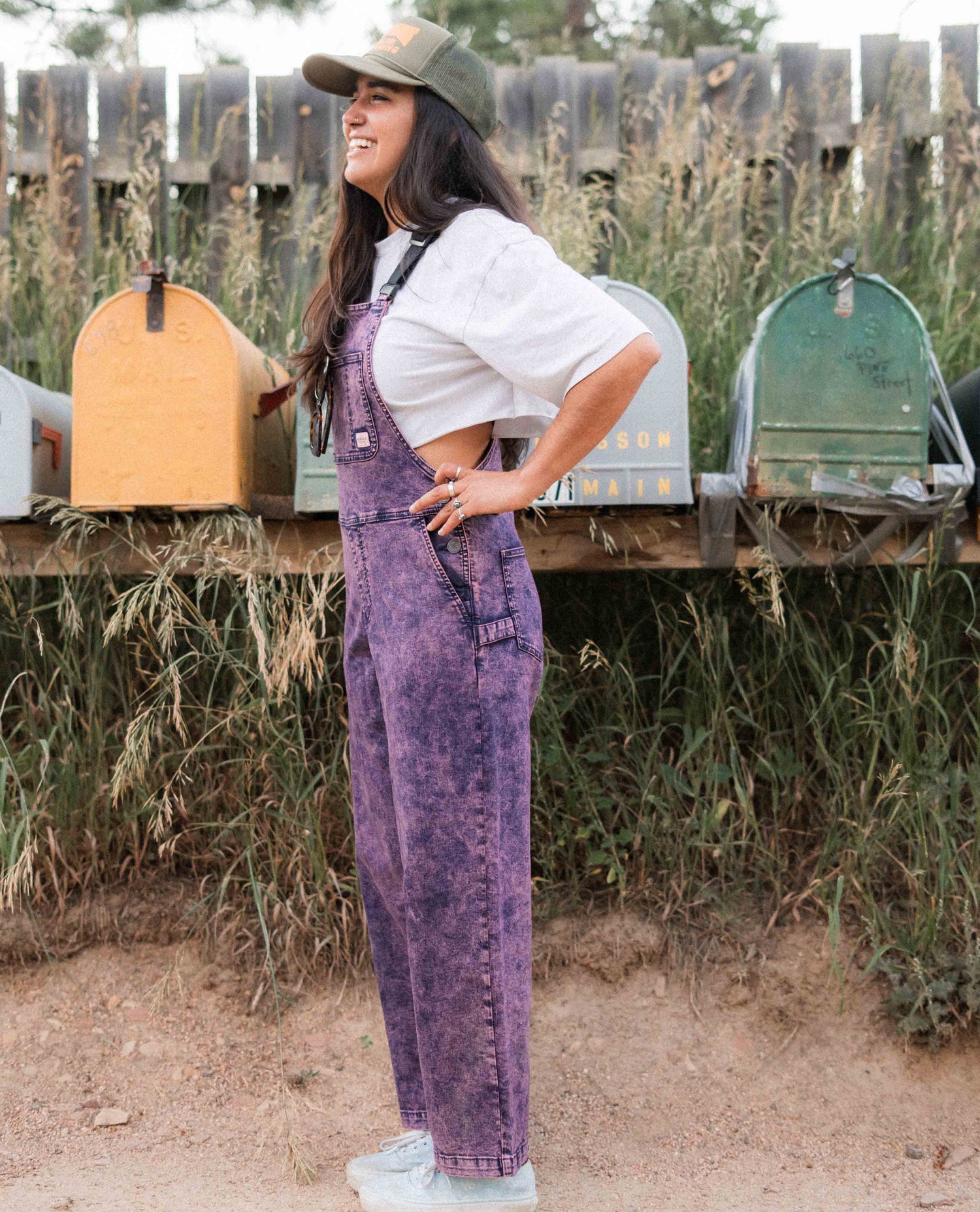 Person wearing purple acid-wash overalls standing in front of a wooden fence with mailboxes.