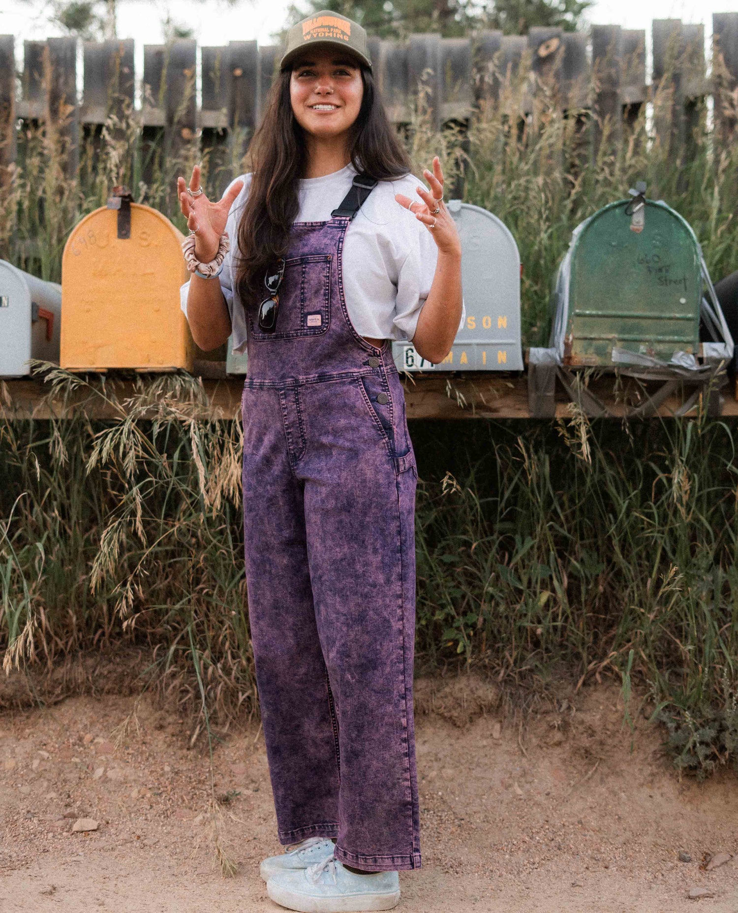 Person wearing purple overalls and a cap standing in front of colorful mailboxes.