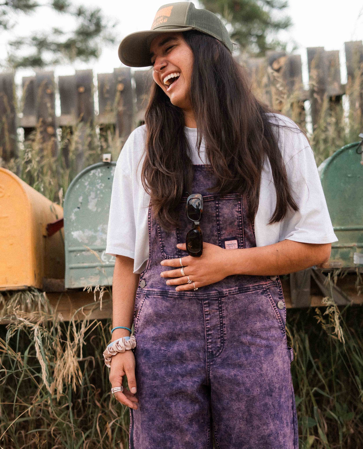 Woman wearing a white t-shirt, purple overalls, and a green cap, standing outdoors with mailboxes in the background.