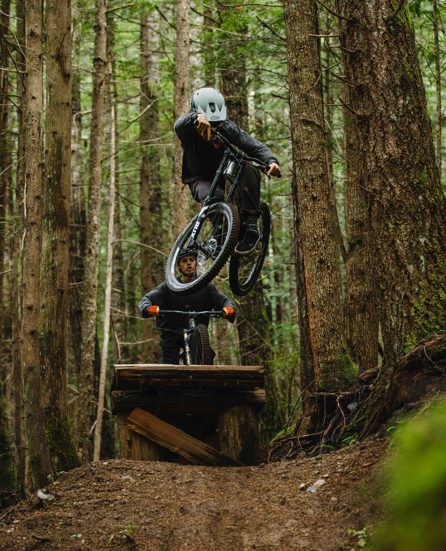 Two mountain bikers in a forest setting with one performing a jump.
