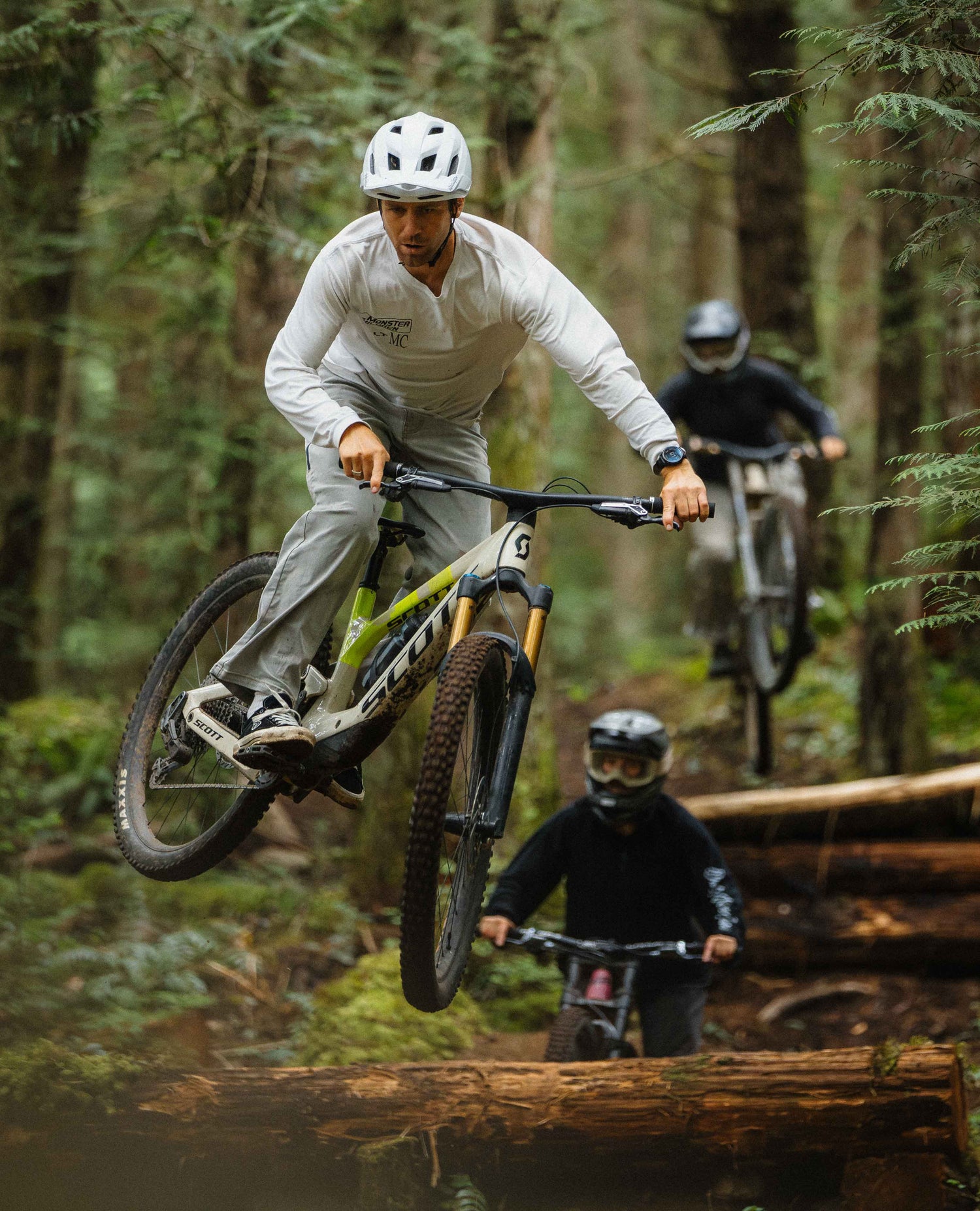 Man riding a mountain bike through a forest with another rider in the background.