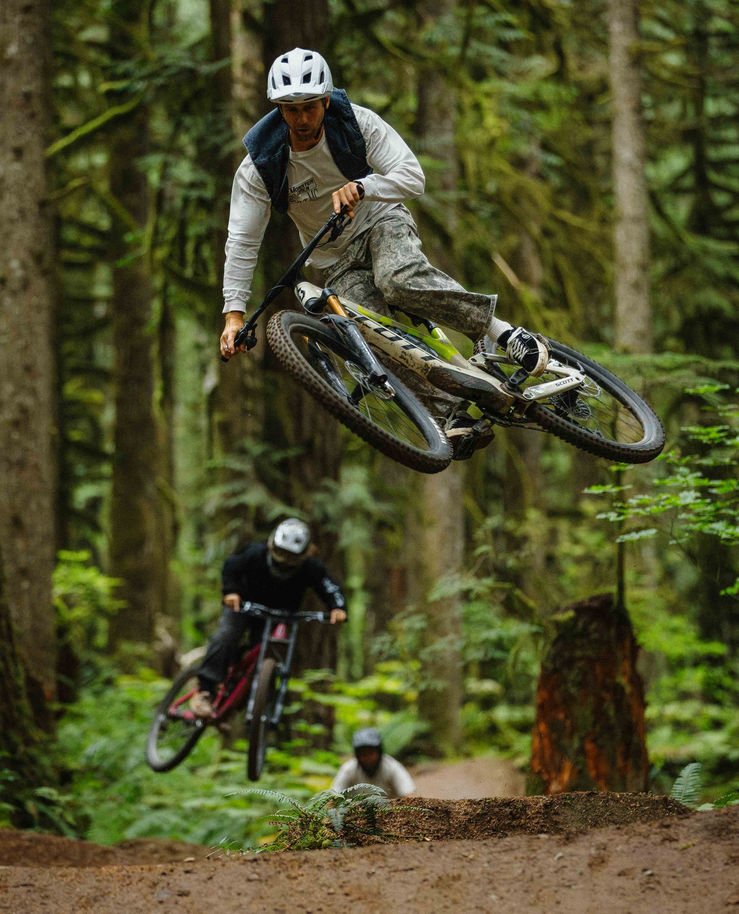 Person riding a mountain bike through a forest with another rider in the background.