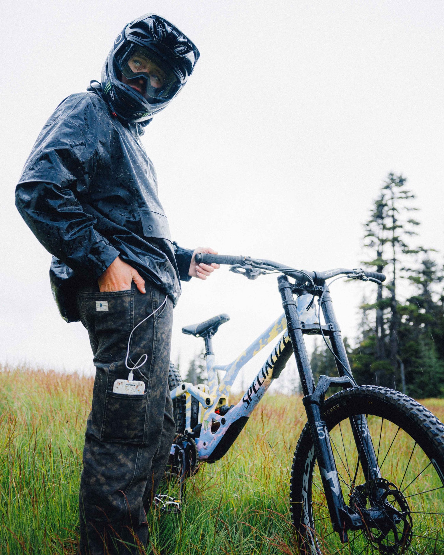 A person in a black rain jacket, helmet, and cargo pants stands beside a mountain bike in an outdoor grassy field under an overcast sky.
