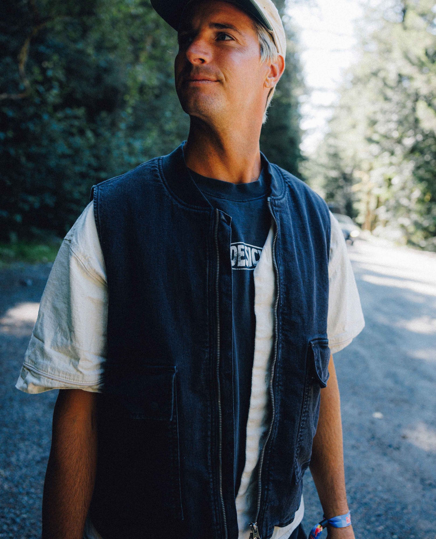 A man wearing a dark denim vest over a white t-shirt and a beige cap, standing on a sunlit gravel road surrounded by trees.