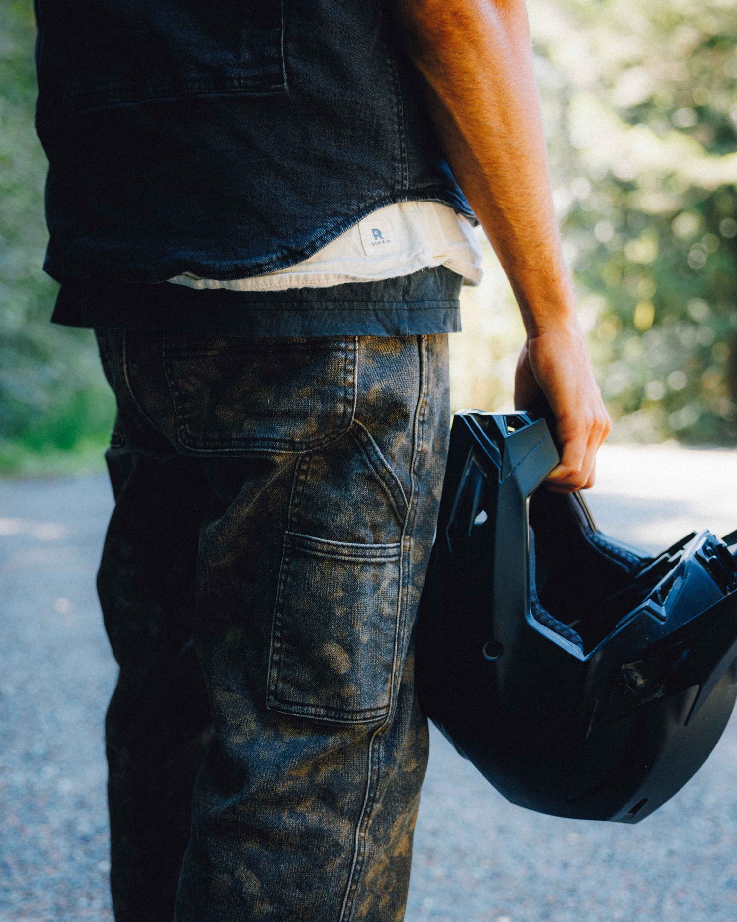 A person outdoors wearing dark washed cargo-style jeans and a dark shirt, holding a black motorcycle helmet in one hand.