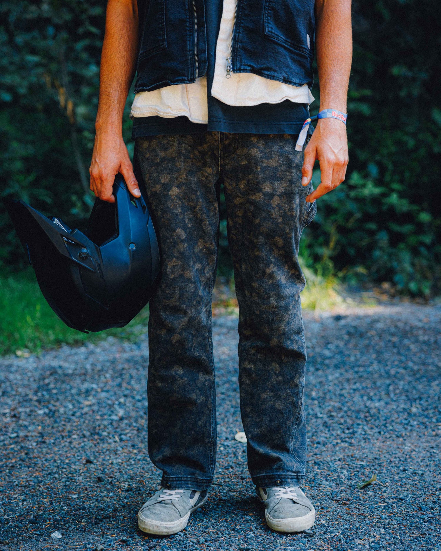 A person standing outdoors on a gravel path wearing dark washed denim jeans, a layered denim vest and white shirt, holding a black motorcycle helmet.