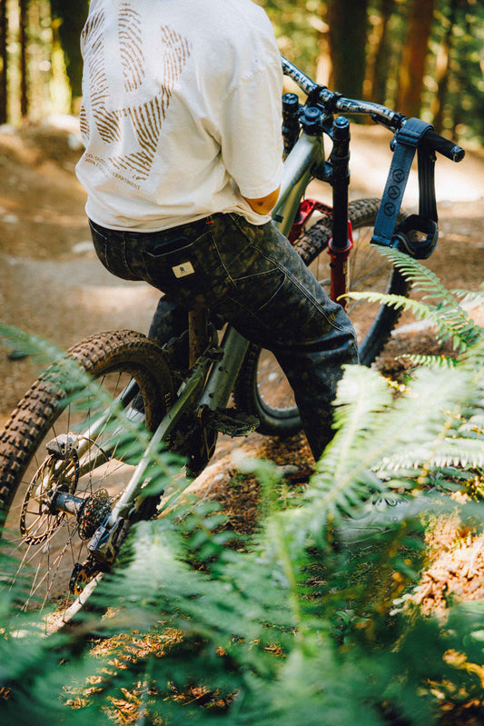 A person riding a mountain bike through a forest trail, viewed from behind. The rider wears a white t-shirt with a gold graphic on the back and dark jeans, with green ferns in the foreground and trees in the background.