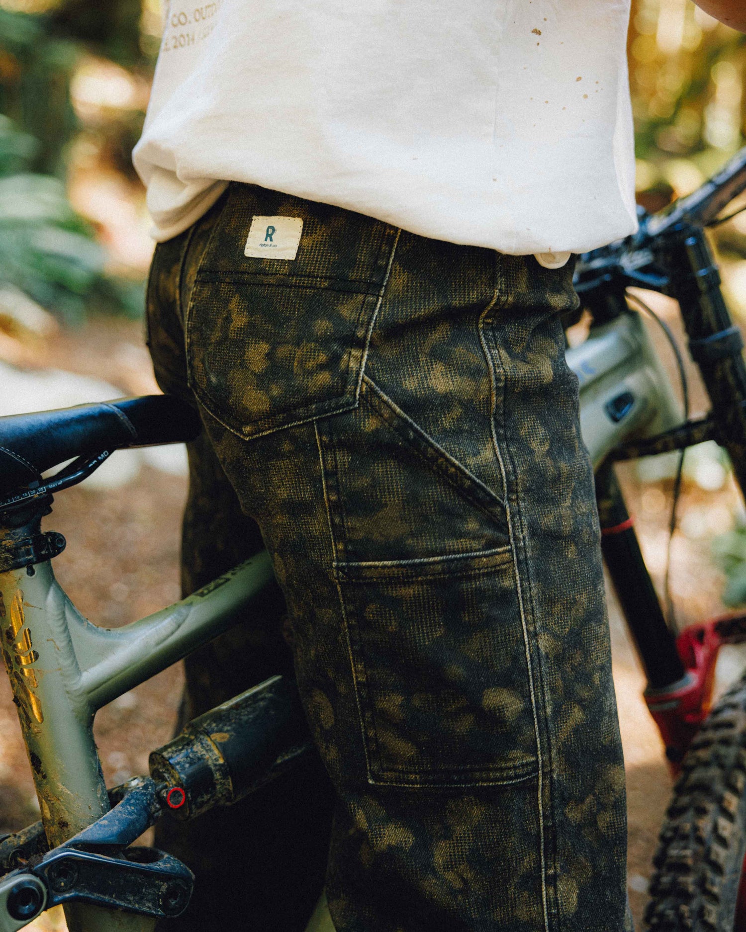 Close-up of a person wearing dark olive camouflage work pants and a white t-shirt, standing next to a mountain bike in an outdoor forest setting.