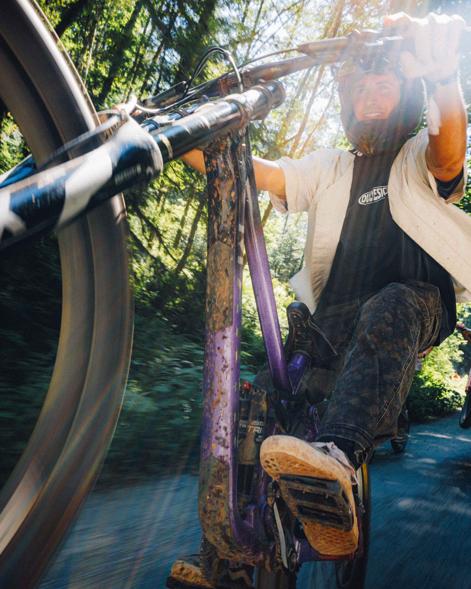 A mountain biker wearing a helmet and light jacket rides a purple bike on a forest road, with the front wheel and rider's legs visible in a dynamic action shot.