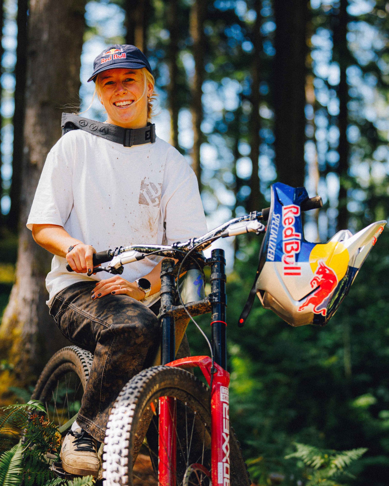 A smiling woman wearing a Red Bull cap and a white sponsor jersey sits on a red mountain bike in a forest. A Red Bull helmet rests on the handlebar area, with a blurred background of trees and foliage.