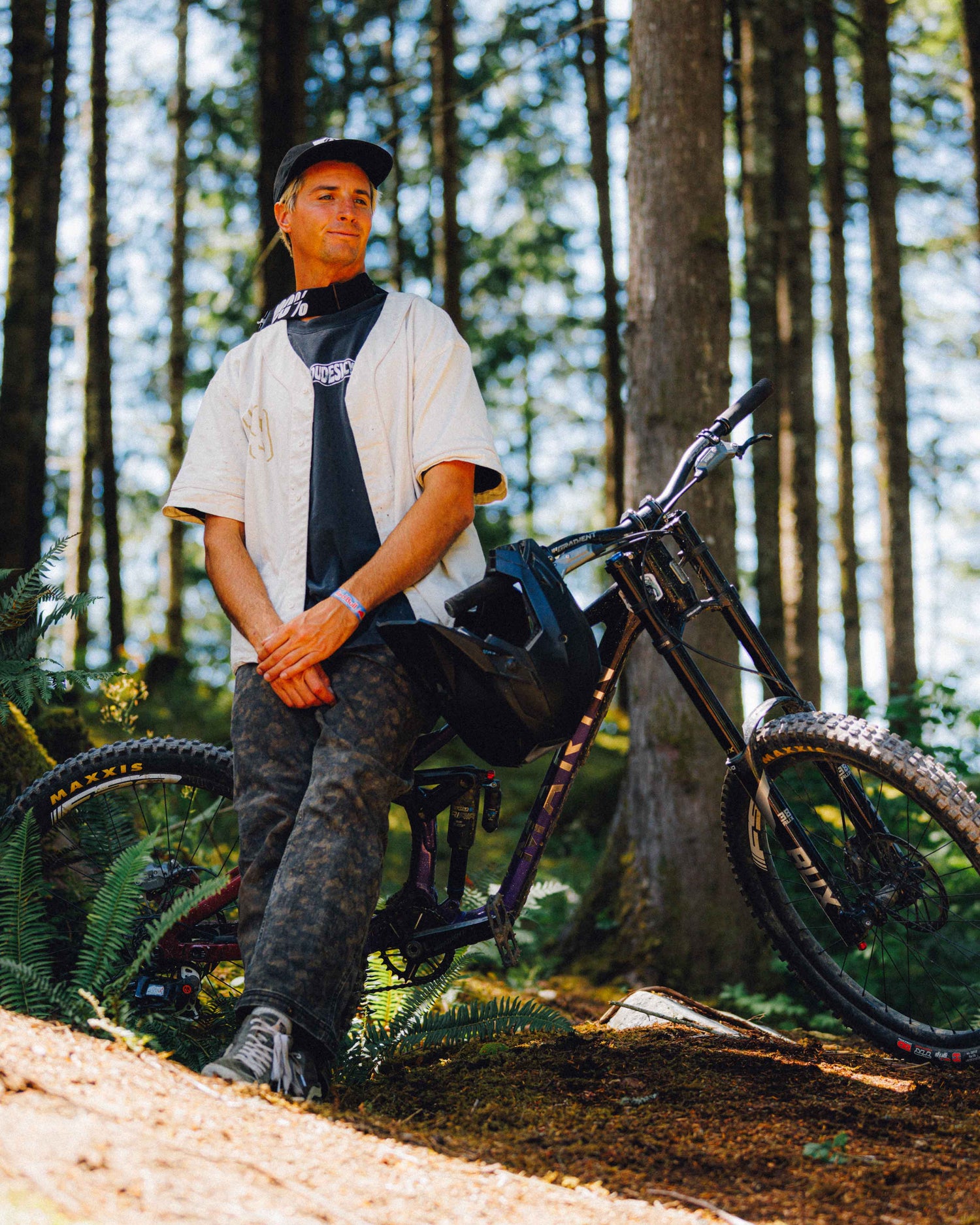 A man wearing a black cap, white short-sleeve shirt, and camouflage pants sits on a black mountain bike in a sunlit forest with tall trees and ferns.