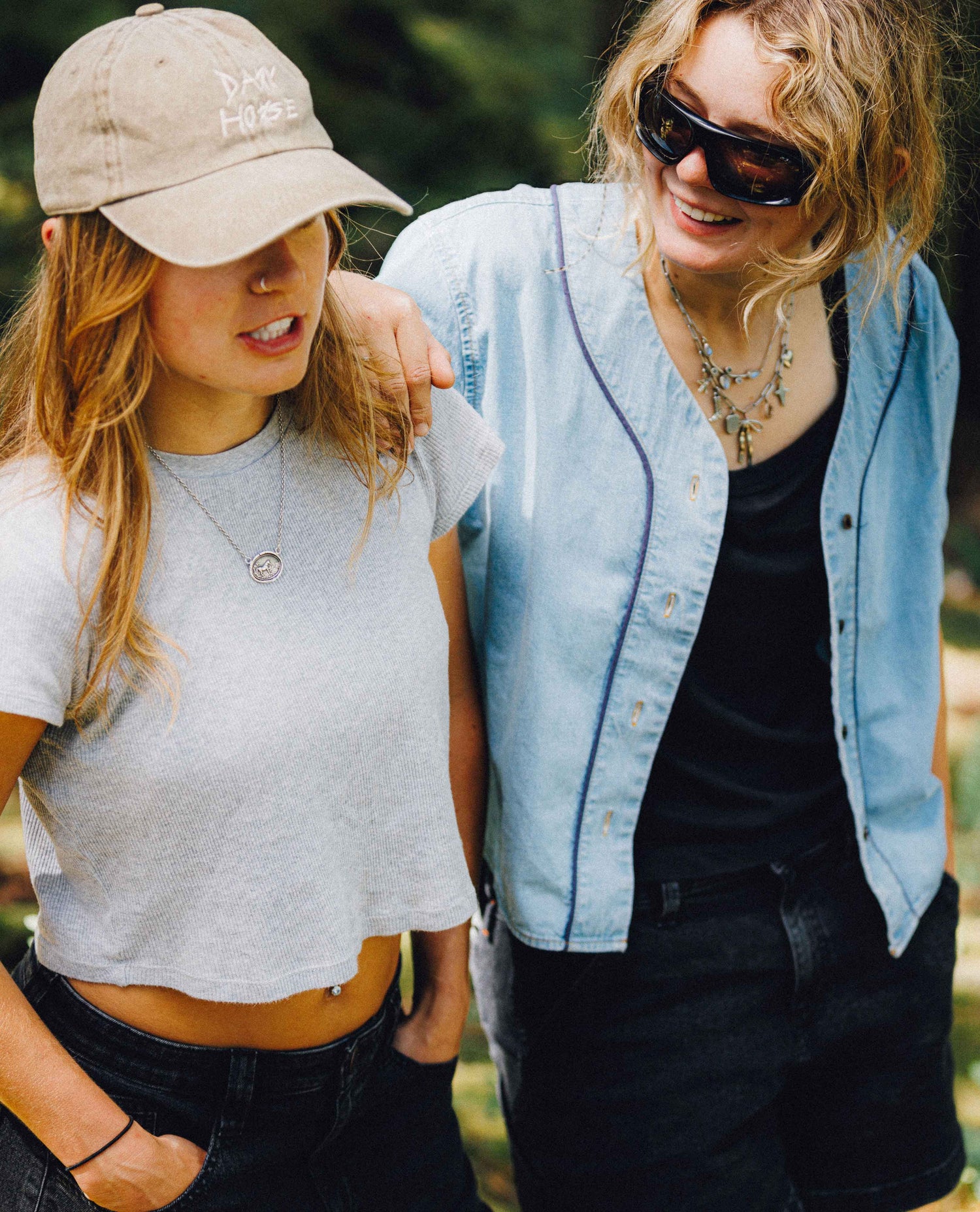 Two women standing outdoors in a wooded setting. The woman on the left wears a beige baseball cap with white embroidered text, a light gray cropped t-shirt, and dark jeans.