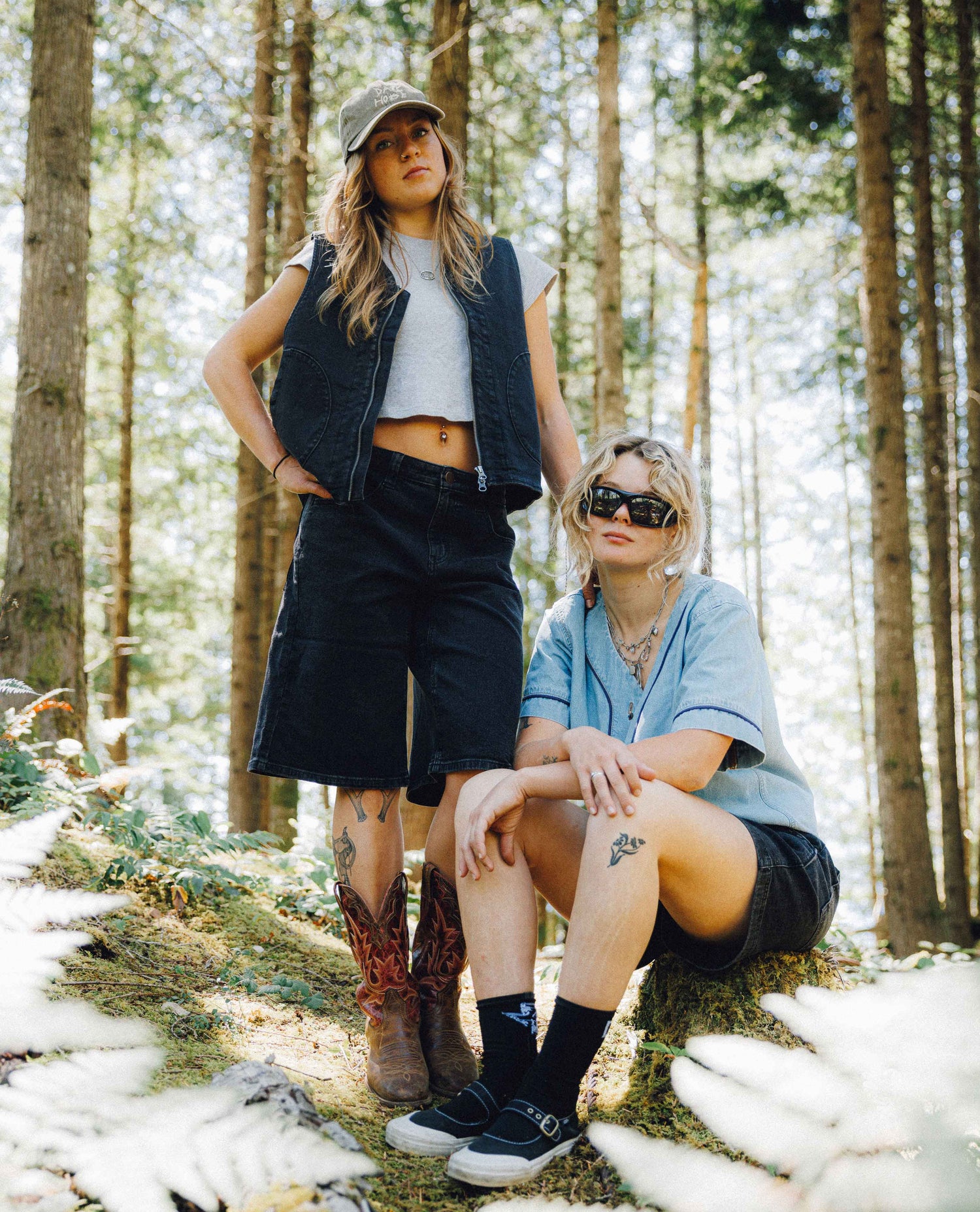 Two women posing in a sun-dappled forest. The standing woman wears a gray cap, a dark denim vest over a white crop top, dark denim shorts, and brown cowboy boots.