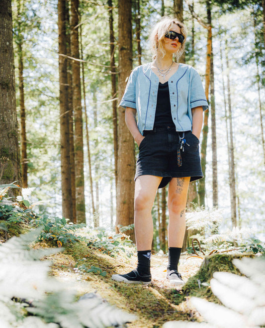 A woman stands in a sunlit forest wearing a light blue cropped button-front shirt, a black mini skirt, a black top underneath, black socks, and black sneakers.