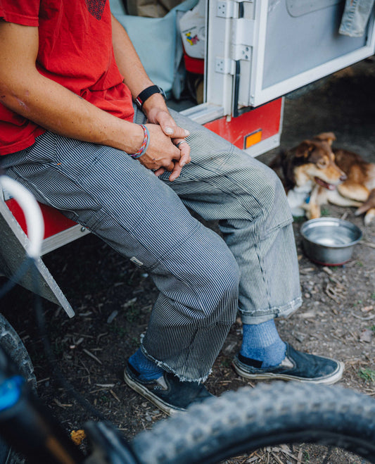 A person sitting on the edge of a red and white utility vehicle outdoors, wearing a red t-shirt, gray pinstriped cargo pants, blue socks, and dark slip-on shoes. A brown dog is lying on the ground near a metal water bowl in the background.