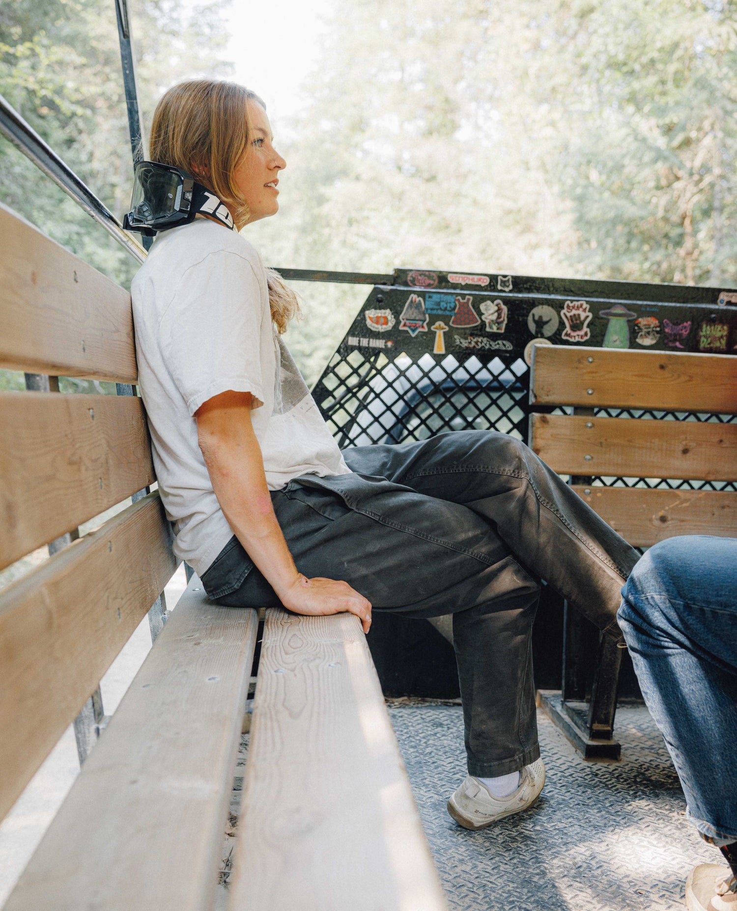 A woman sits on a wooden bench inside a vehicle, wearing a white t-shirt, dark gray jeans, and white sneakers. The background shows trees and a vehicle interior with stickers.