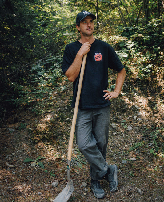 A man standing on a wooded trail holding a shovel, wearing a black t-shirt with a red and white graphic on the left chest, gray pants, and a black cap.