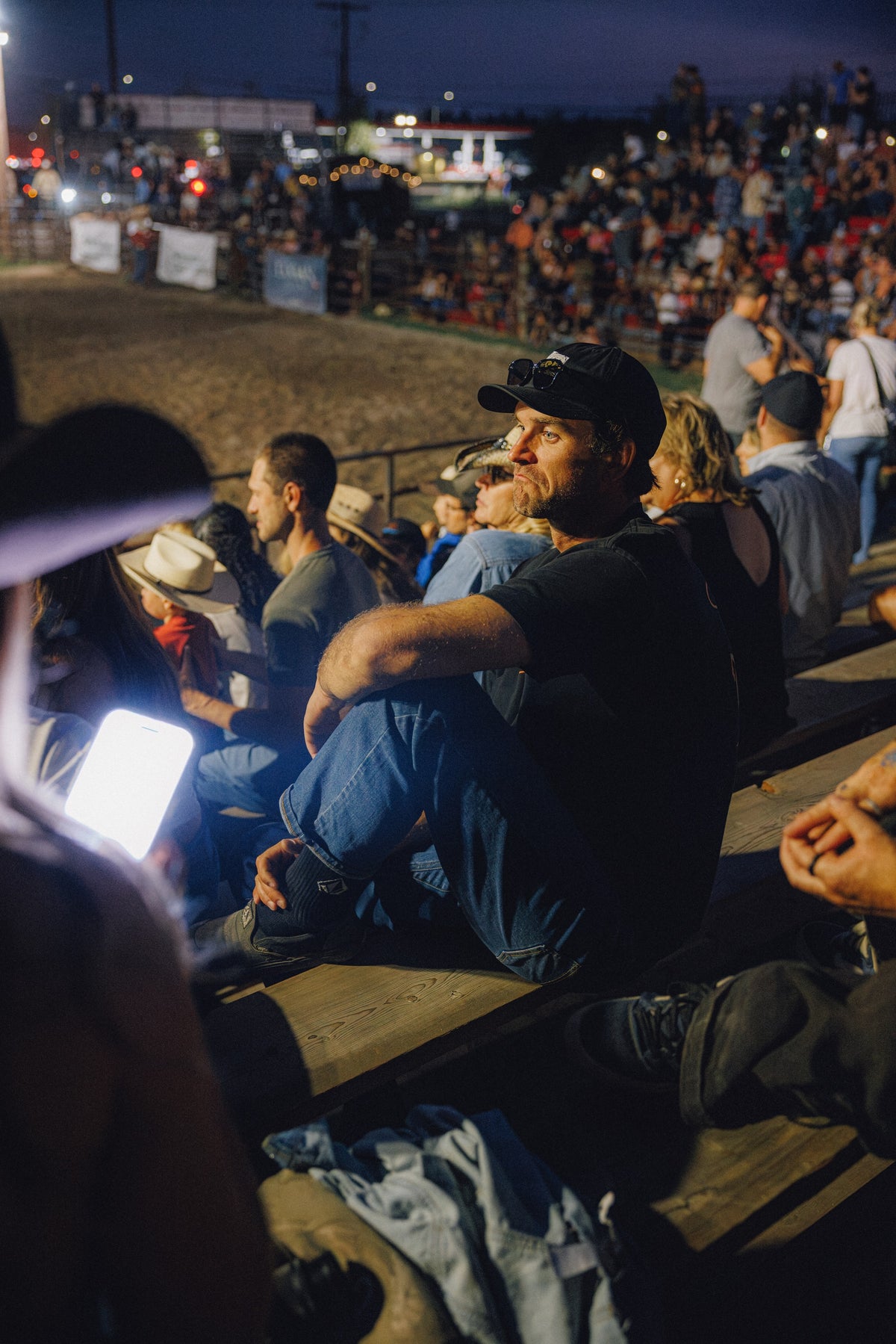 A man in a black cap, black t-shirt, and blue jeans sits in the stands at a night event or rodeo, looking off-camera with a phone in his lap. The background shows a crowd of spectators and arena lights.