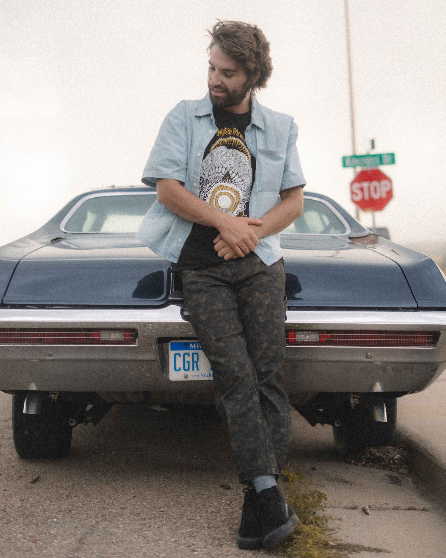 A man leans against the rear of a dark blue classic car outdoors, wearing a light blue button-up shirt over a black graphic t-shirt, dark camouflage pants, and black shoes.