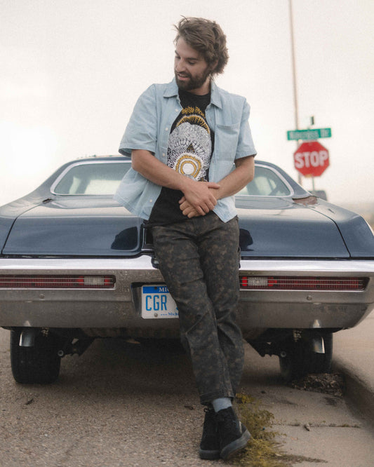 A man leans against the rear of a dark blue classic car outdoors, wearing a light blue button-up shirt over a black graphic t-shirt, dark camouflage pants, and black shoes.