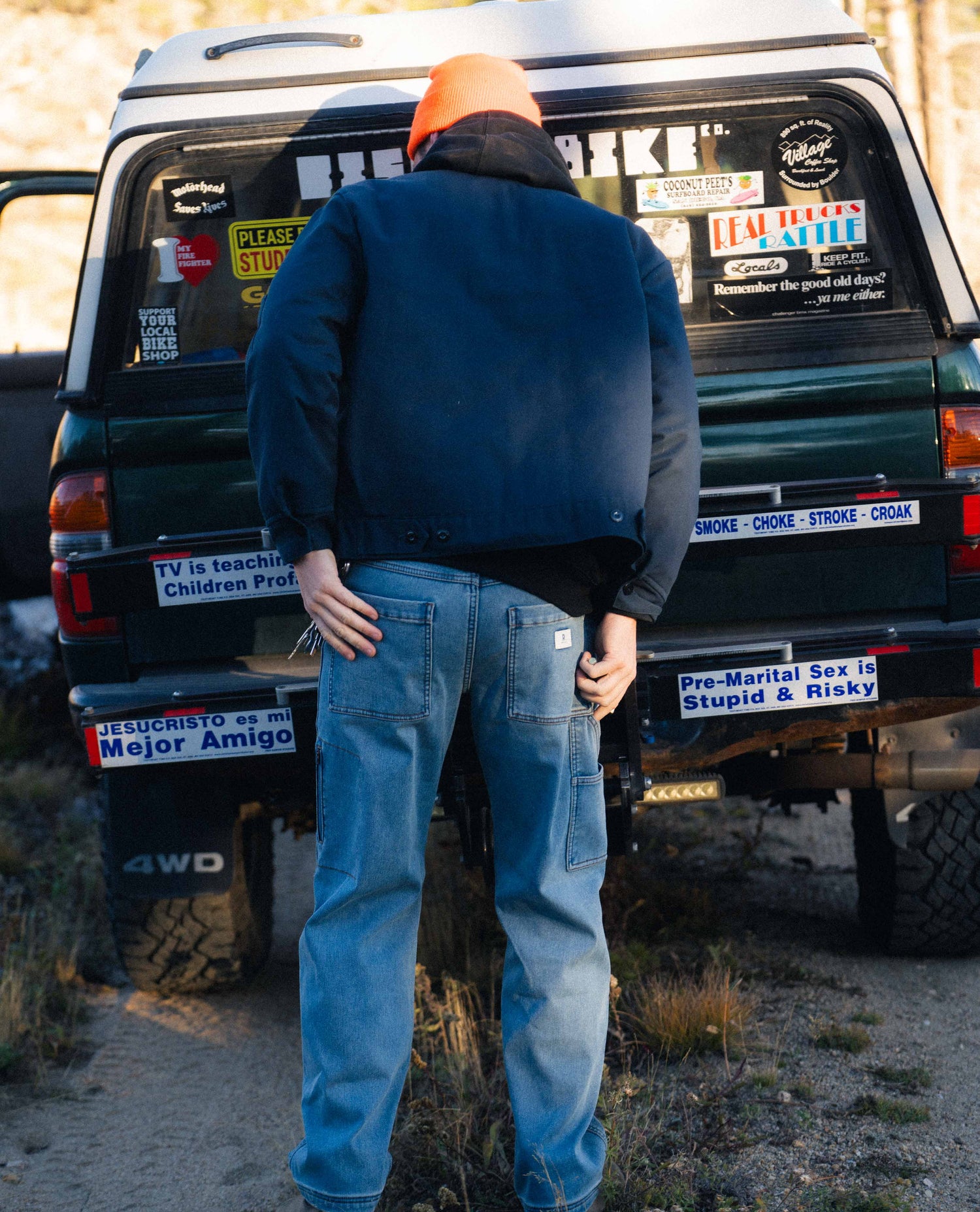 Person standing next to a green SUV with various stickers on a dirt road