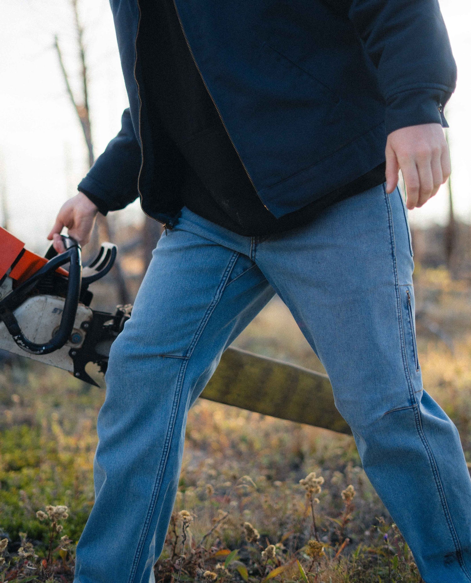 Person wearing blue jeans and a dark jacket holding a chainsaw in an outdoor setting.