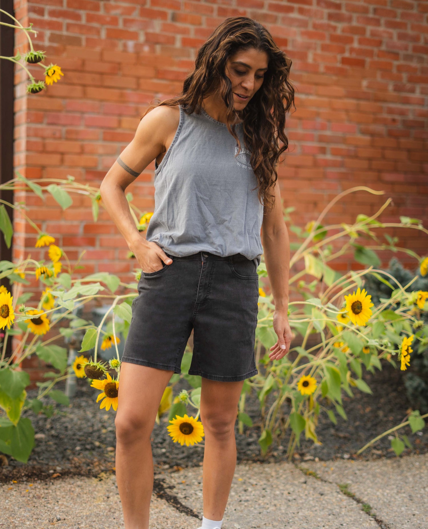 Woman in gray tank top and black shorts standing in front of a brick wall with sunflowers.