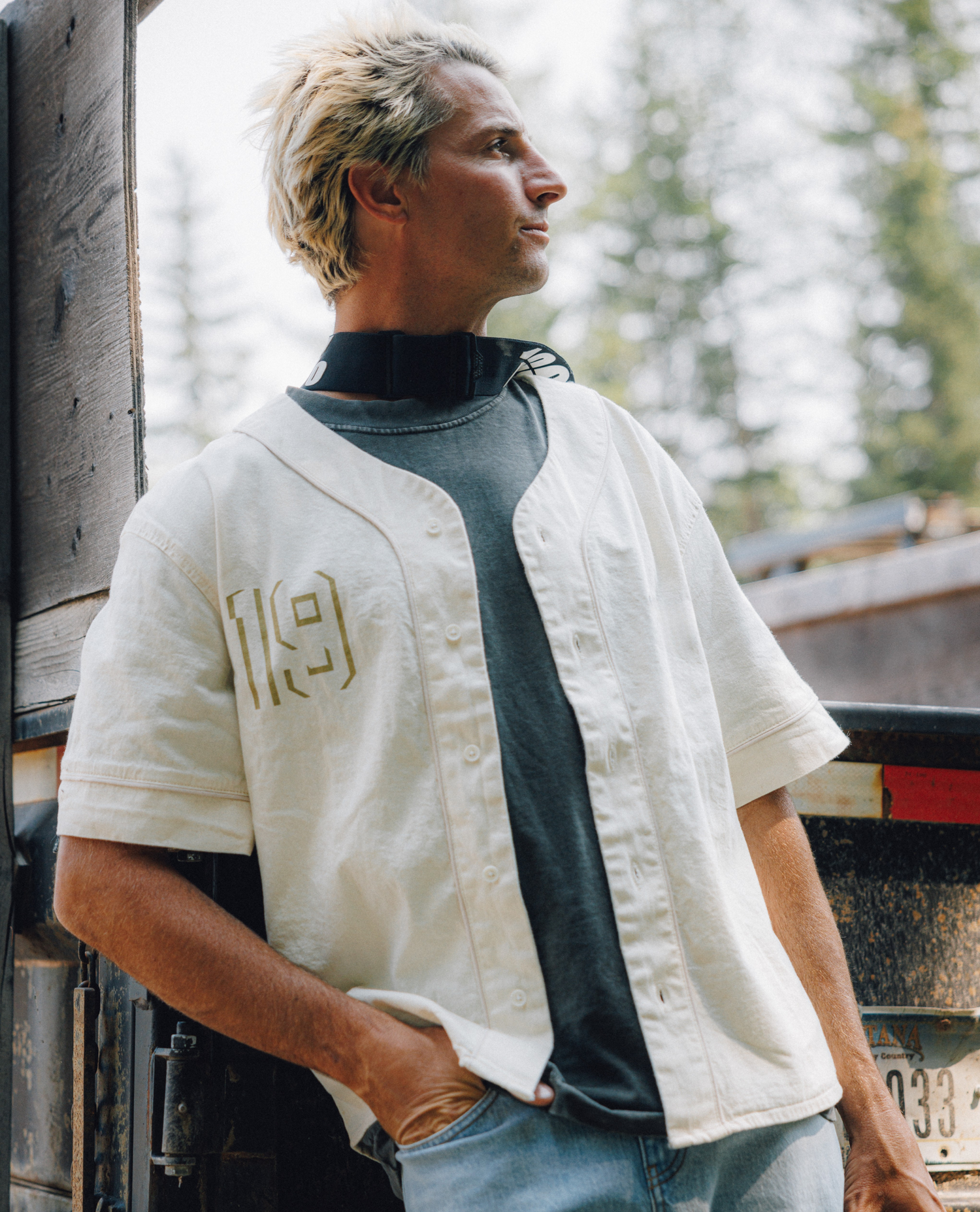 A man wearing a cream-colored short-sleeve baseball-style shirt with gold outlined '19' lettering on the chest, layered over a dark gray mock neck shirt and light blue jeans, standing outdoors next to a rustic wooden structure.