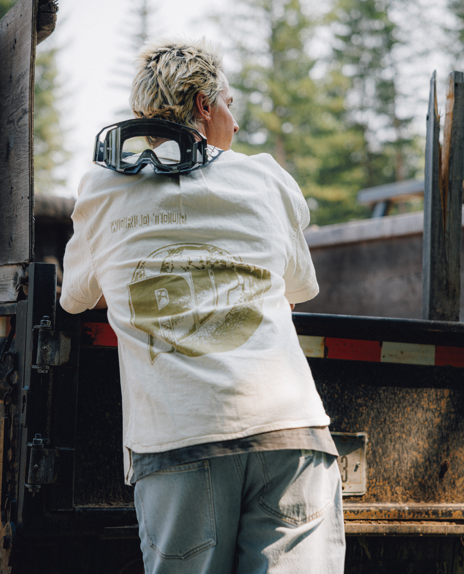 A person seen from behind wearing a cream-colored graphic t-shirt with a large olive-green print on the back, light-wash jeans, and black goggles resting on their neck, standing next to a rusty truck in an outdoor setting.