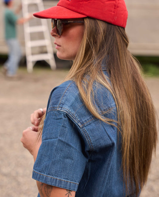 A woman in profile wearing a red baseball cap, dark sunglasses, and a blue denim shirt, photographed outdoors with a blurred background.