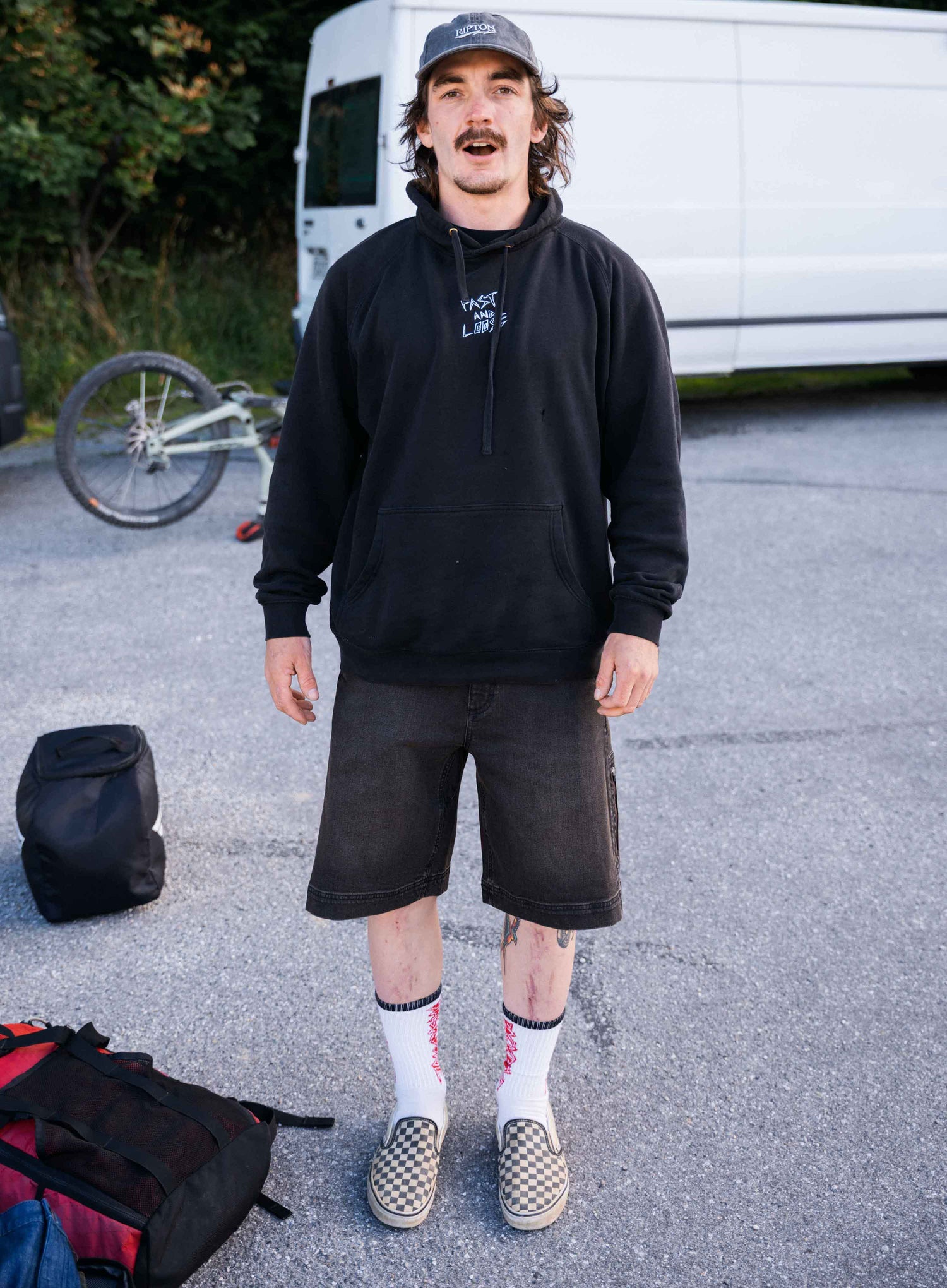 Person wearing a black hoodie and shorts standing in an outdoor setting with a white van and bicycle in the background.