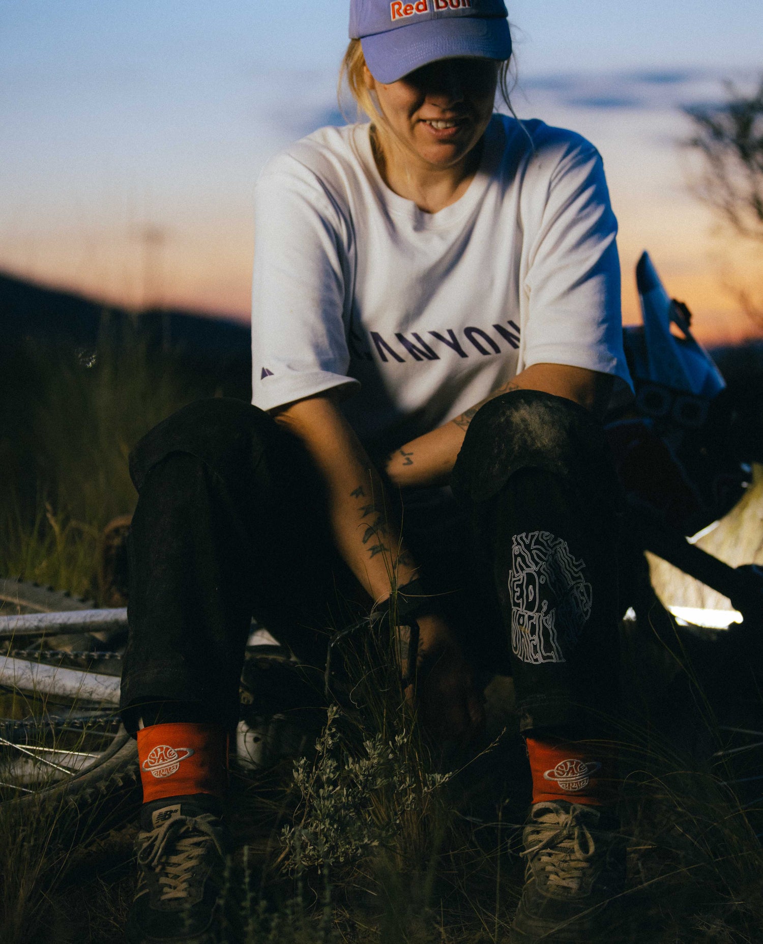 Person sitting outdoors with a scenic background, wearing a white t-shirt and red shoes.