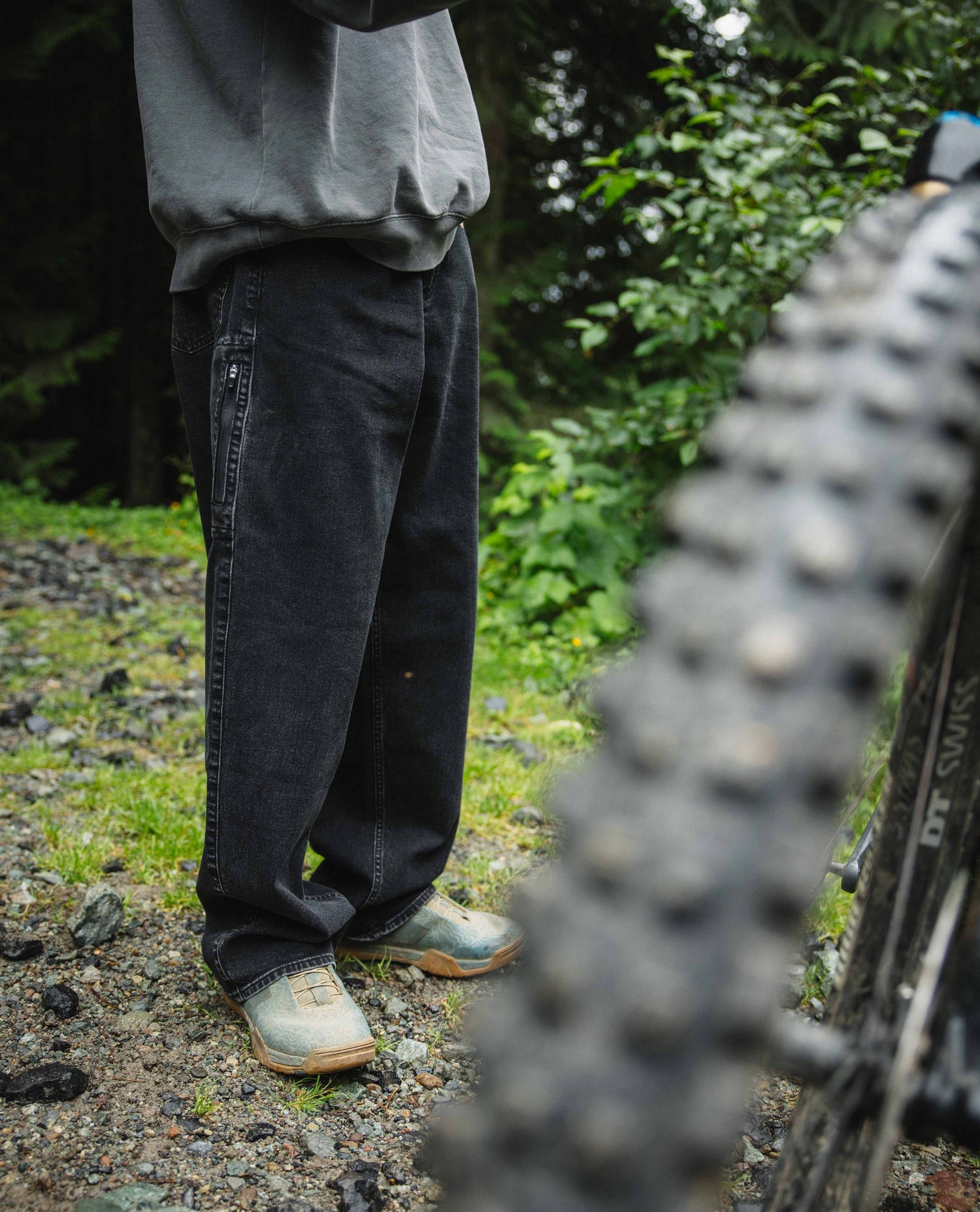Person wearing dark pants and shoes standing next to a bicycle on a trail with greenery in the background