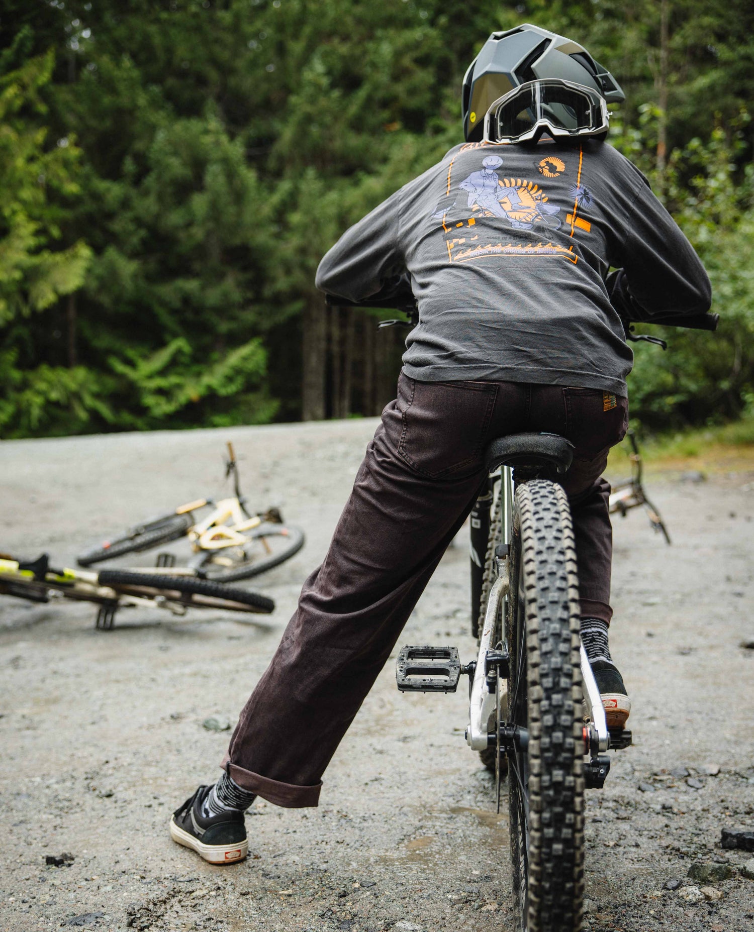 Person on a mountain bike with a helmet and goggles, standing on a dirt path with trees in the background.