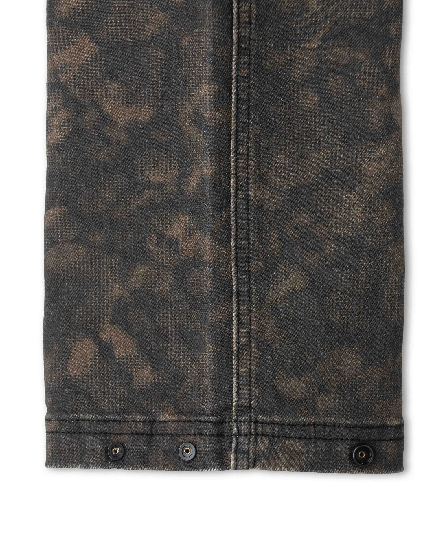 Close-up of a pair of jeans showing the washed dark brown and black leopard print fabric, vertical seam, and buttoned cuffed hem on a white background.