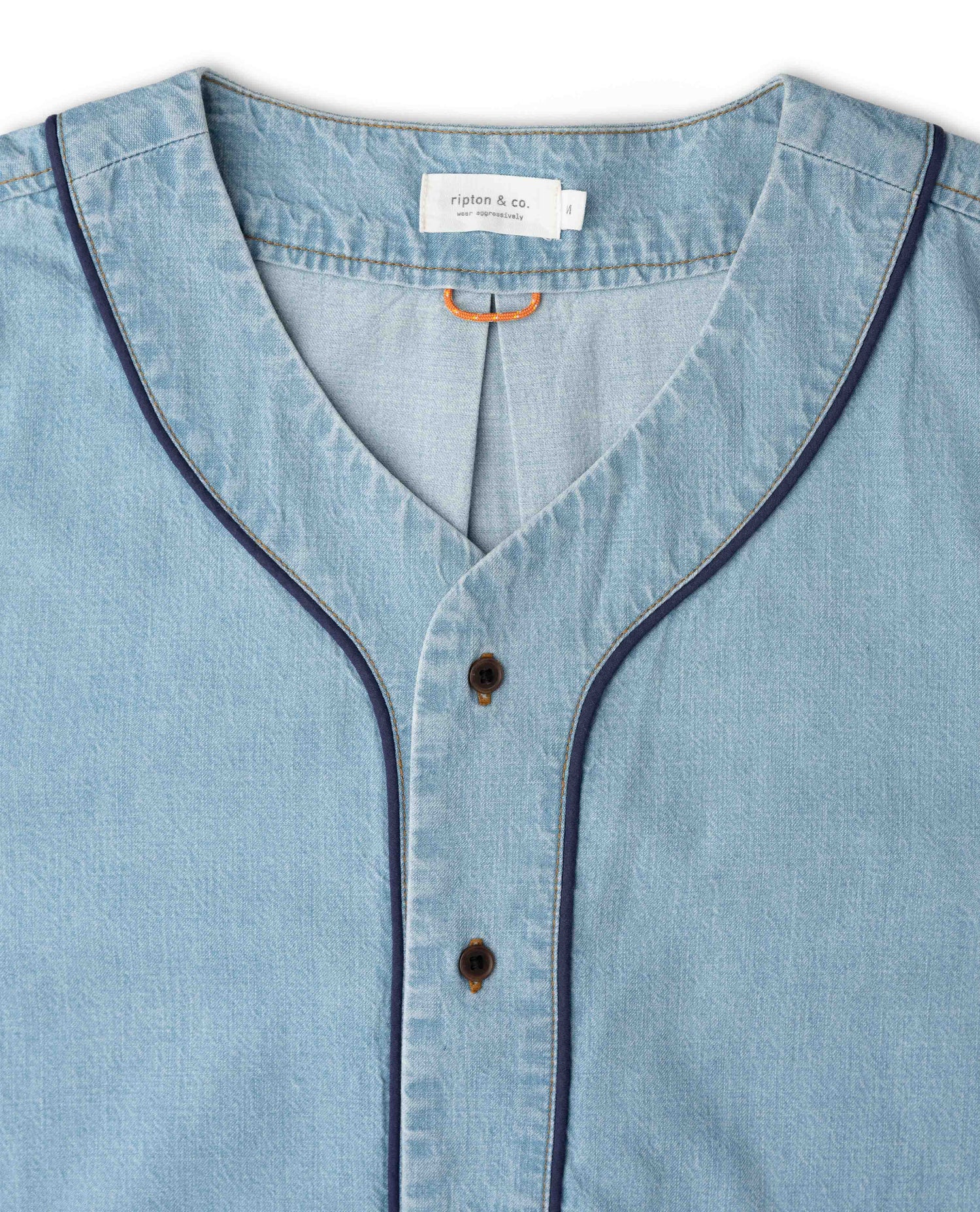 Close-up of a light blue chambray button-up shirt with dark navy piping along the split V-neck and button placket. The brand label 'vintage & co.