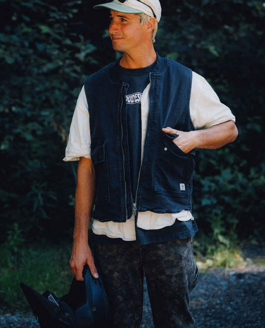 A man wearing a black denim work vest over a white t-shirt and a baseball cap, standing outdoors in a wooded setting.