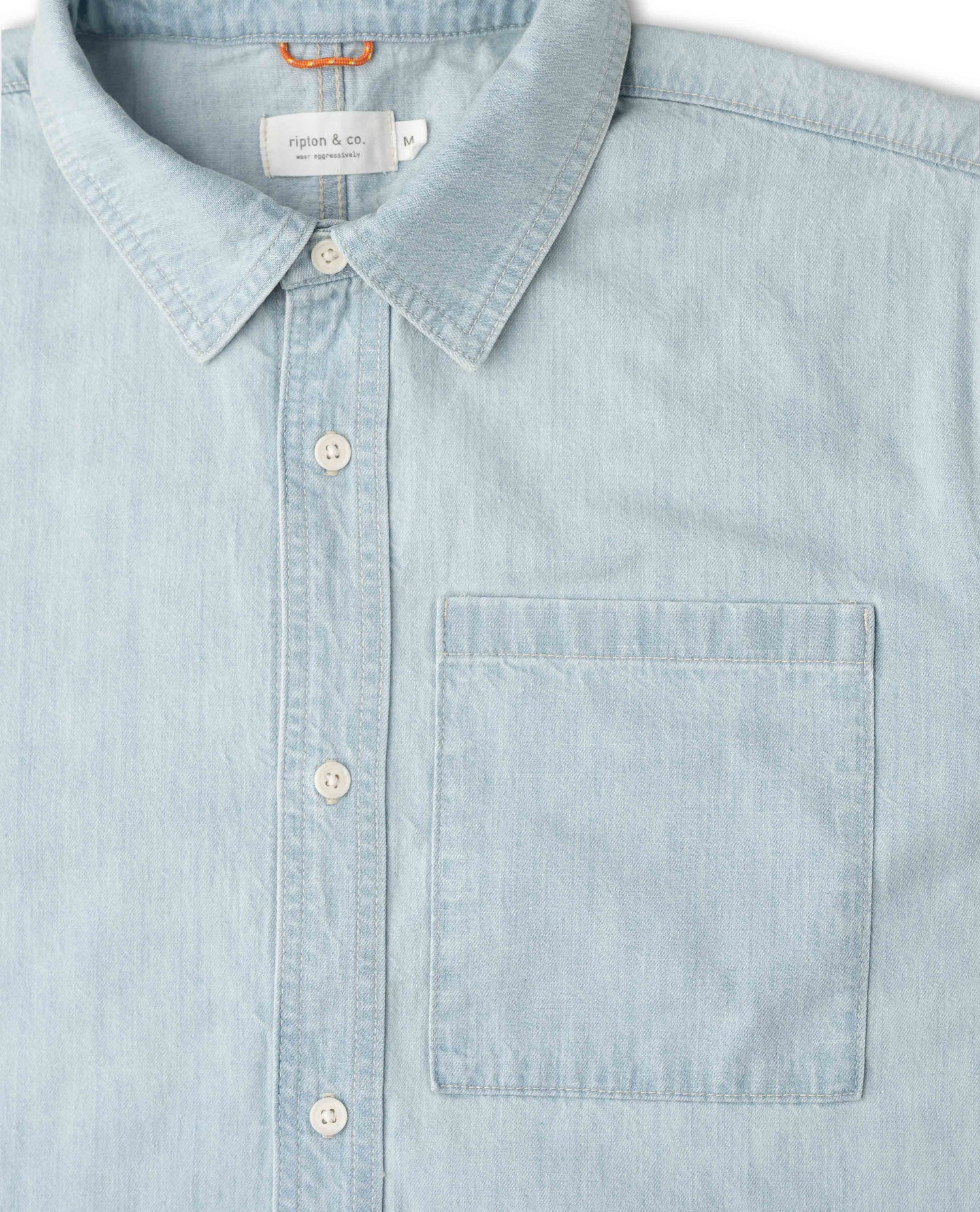 A close-up of a light blue chambray button-down shirt by Ripton & Co, featuring a classic collar, white buttons, and a single chest pocket.