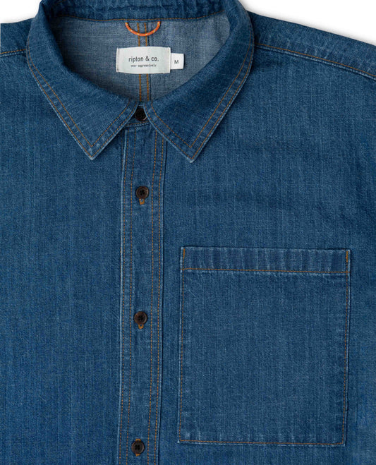 Close-up of a blue denim shirt with a collar and pocket, featuring a brand label.