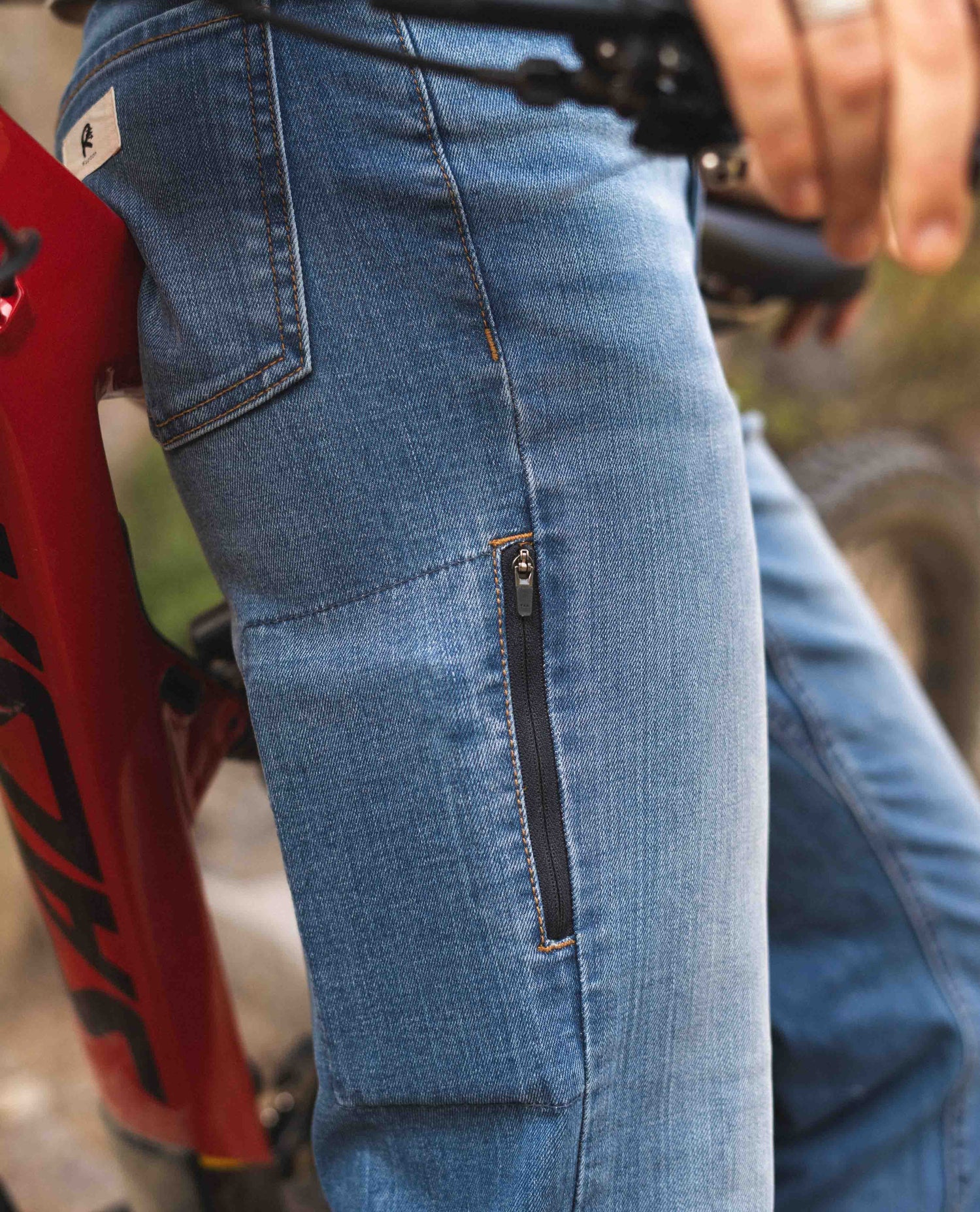 A close up detail of a cell phone pocket on the right leg of a pair of blue jeans. Model is leaning against a red mountain bike.