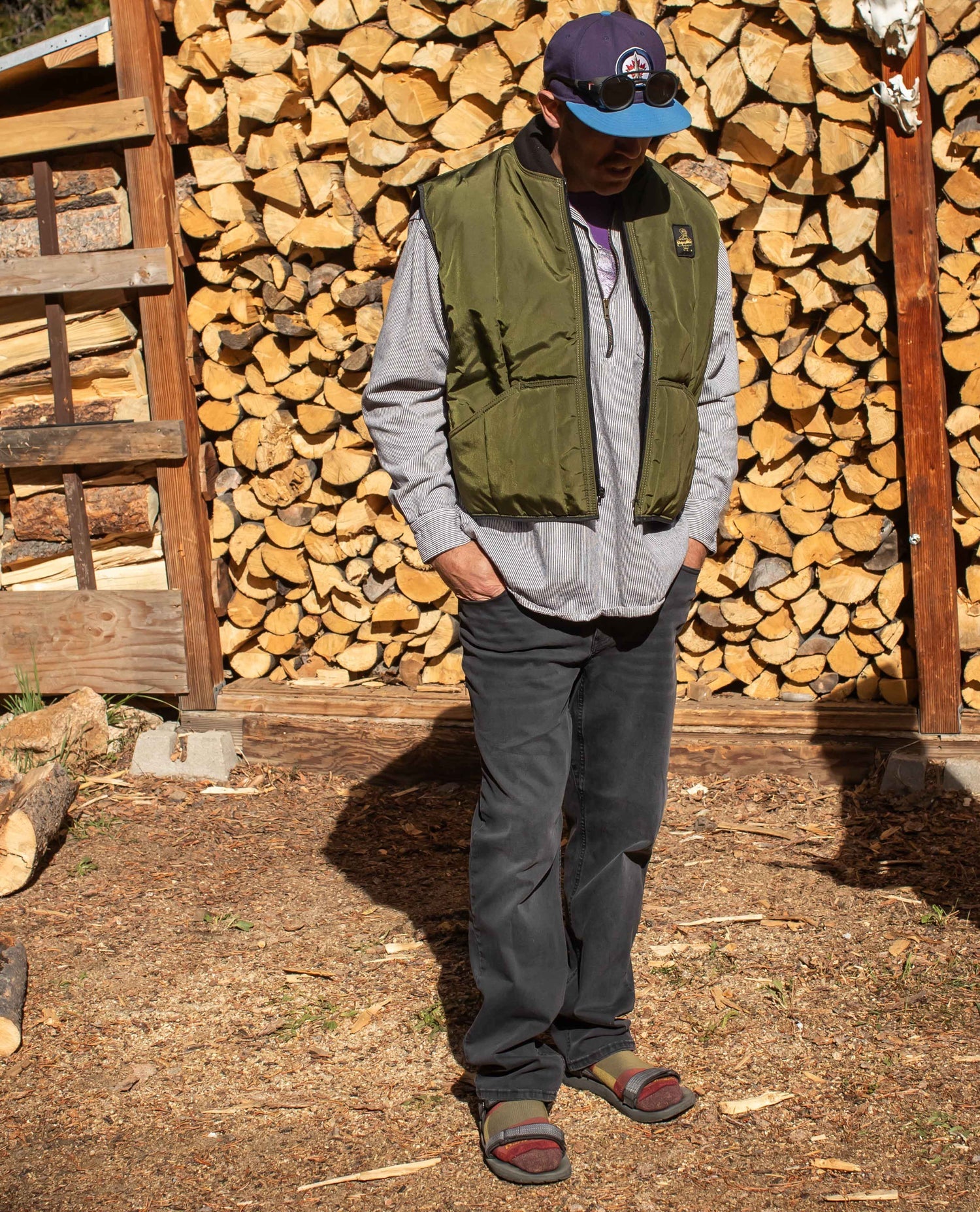 A man standing in front of some firewood. He is wearing a green vest, grey sweatshirt, and black pants. 