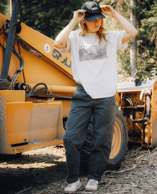 Woman wearing gray hat, gray pants, and white shirt, standing in front of an orange bulldozer.