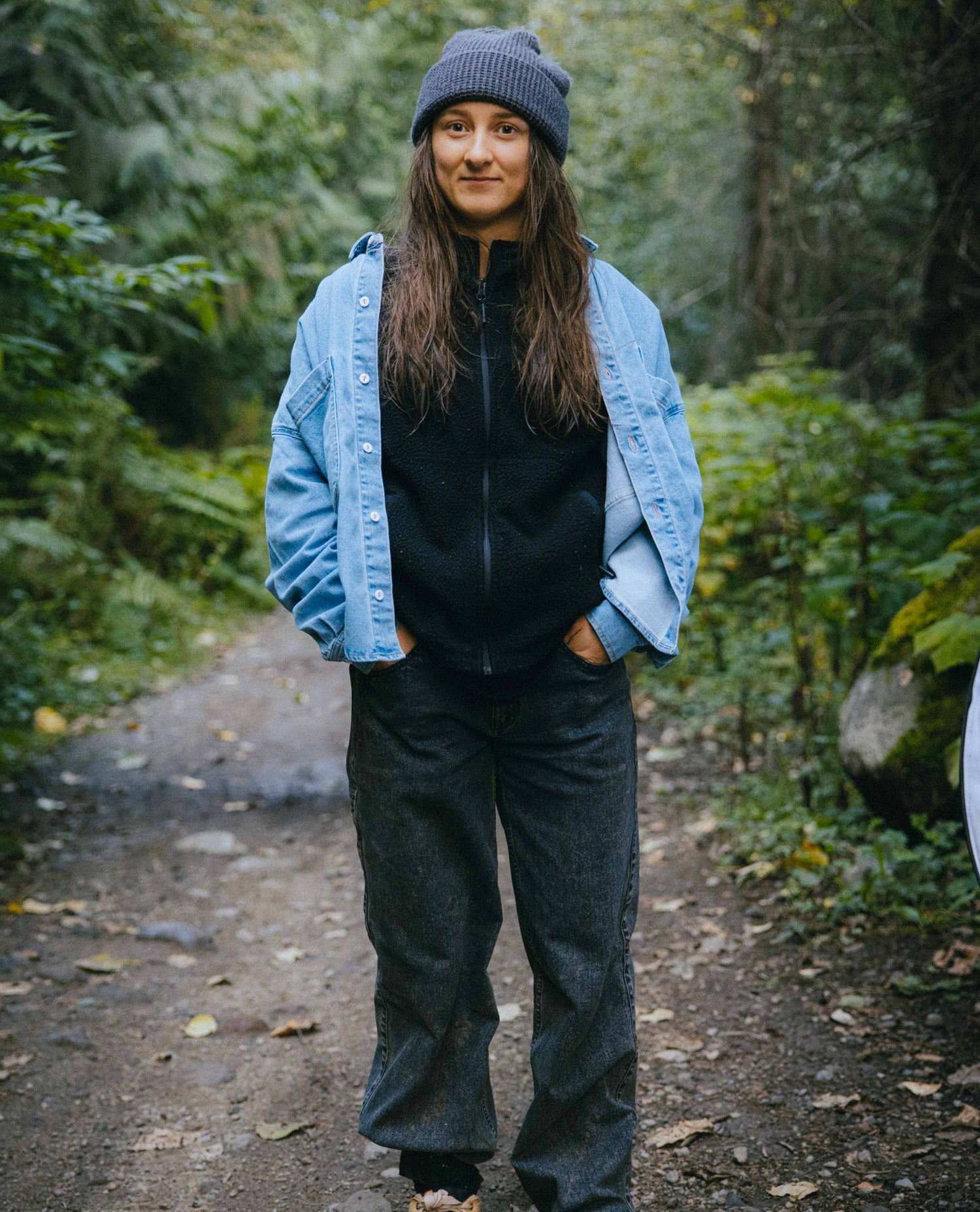 Person standing on a forest path wearing a blue denim jacket, black shirt, and gray pants.