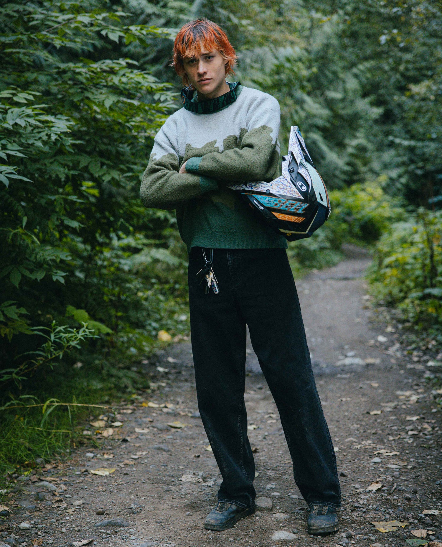 Person with orange hair standing on a forest path holding a helmet