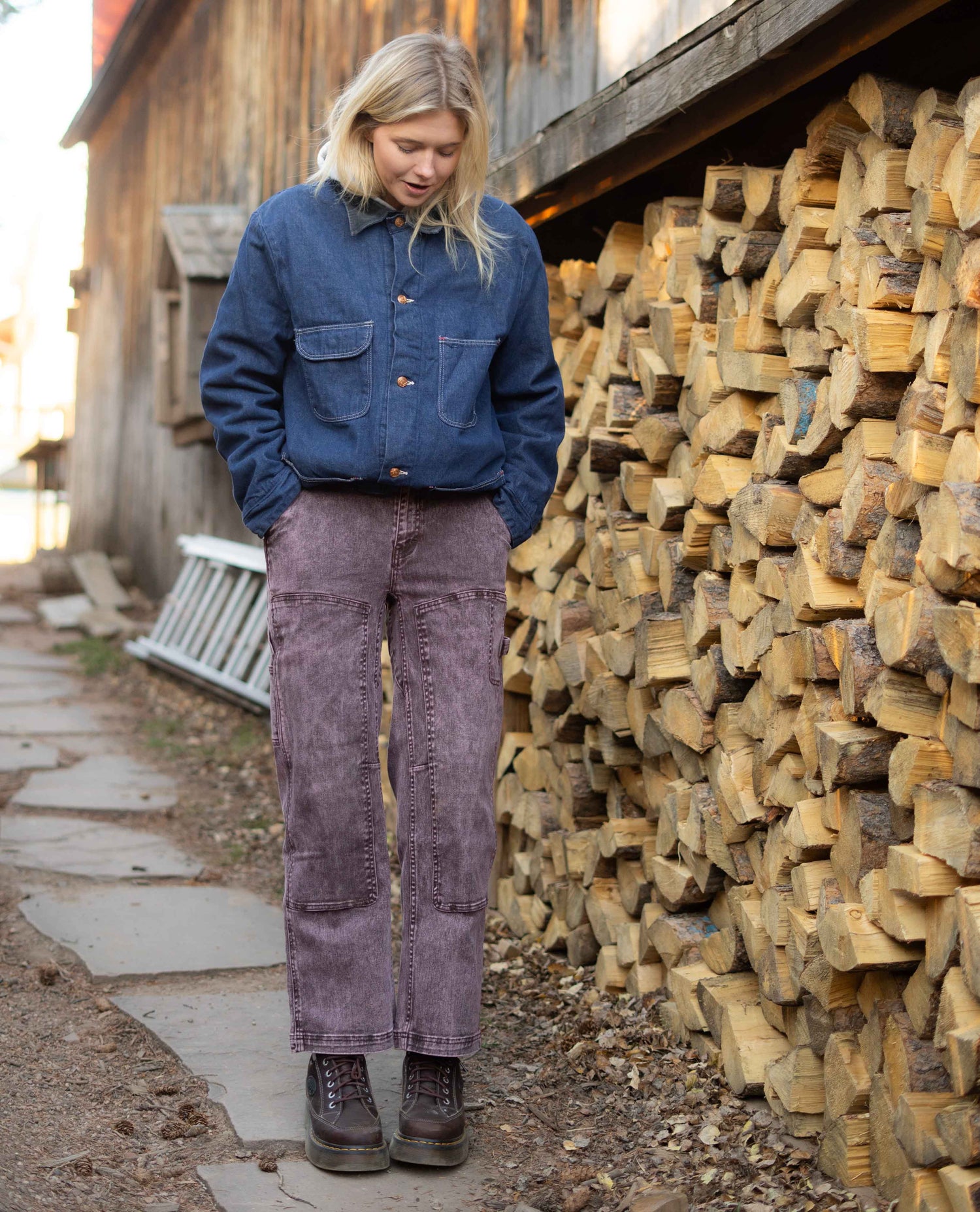 Person wearing a blue denim jacket and purple pants standing next to a stack of wood.