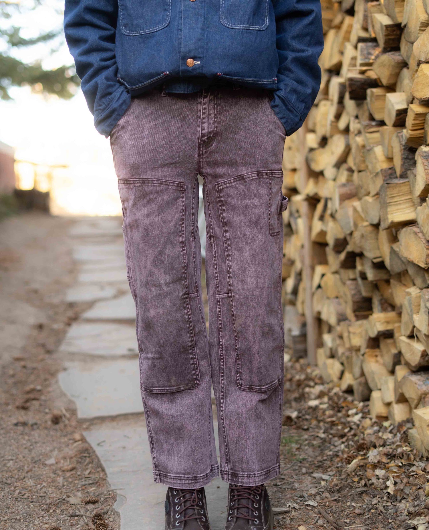 Person wearing a blue jacket and purple pants standing next to a stack of wood.