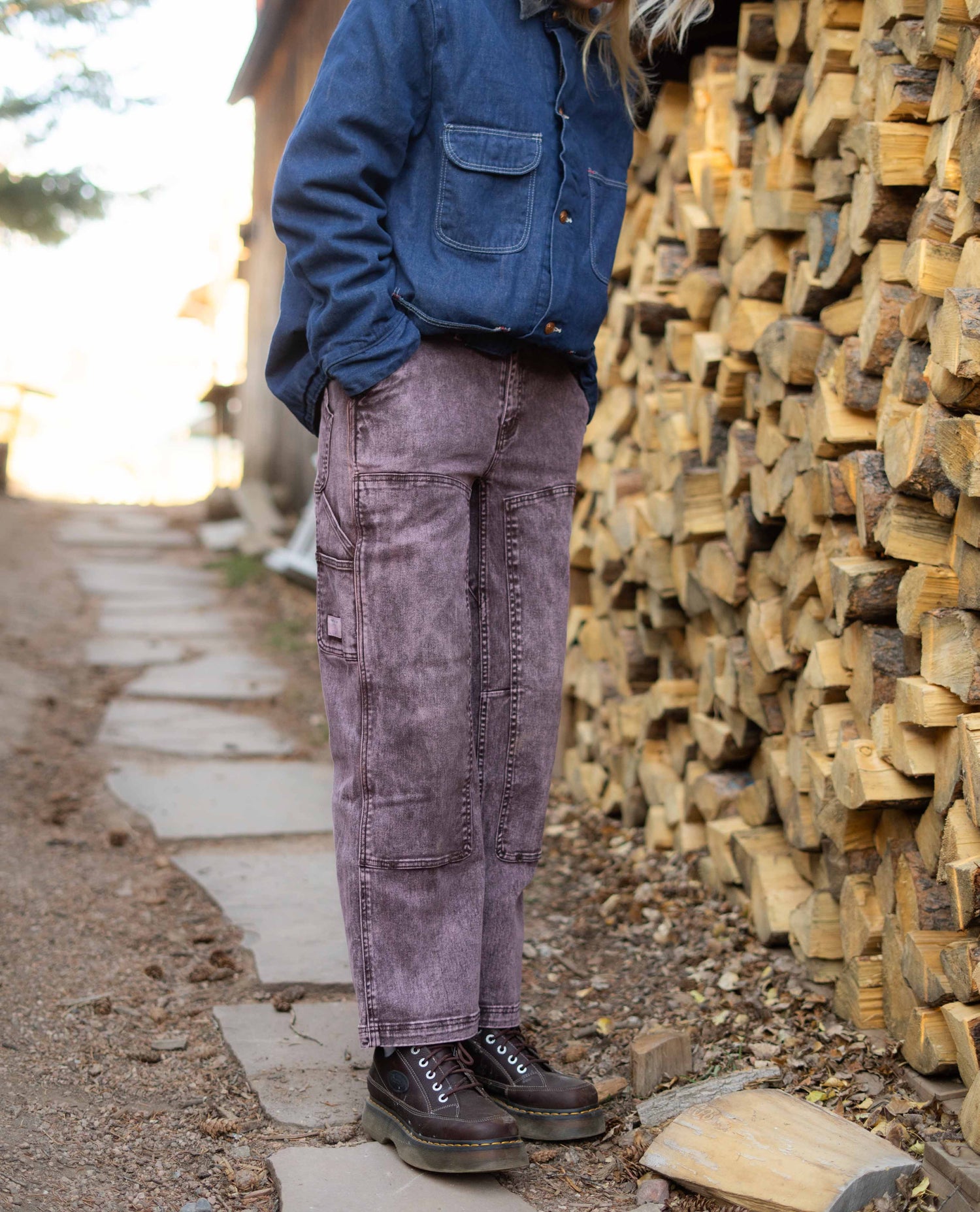 Person wearing a blue jacket and purple pants standing next to stacked firewood.