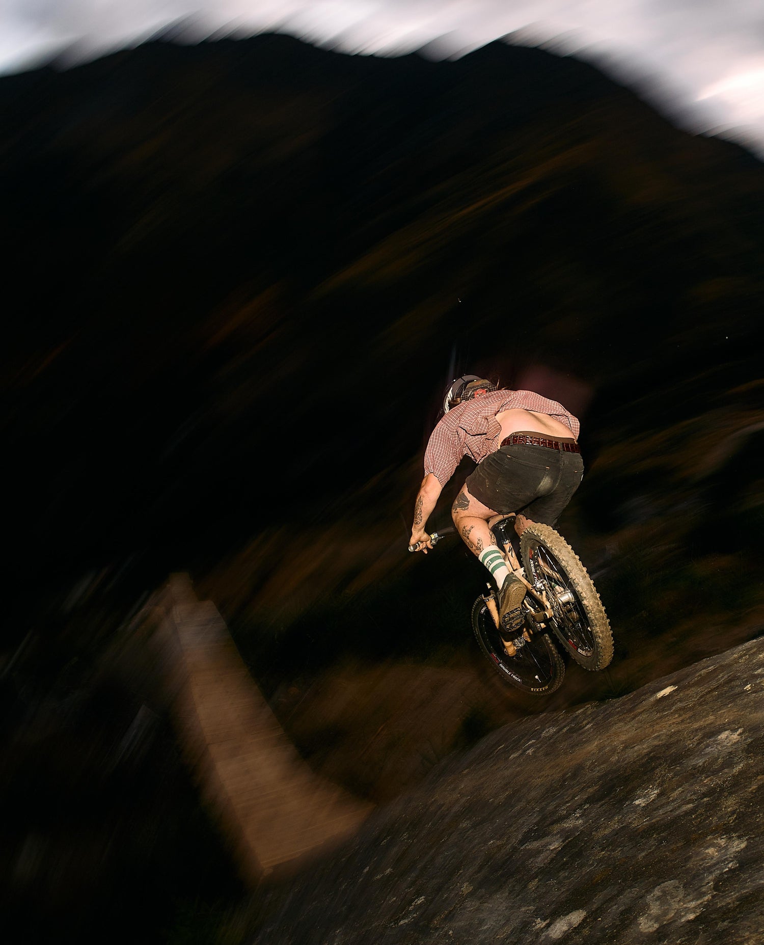 A mountain biker mid-air performing a jump in a dark outdoor setting with motion blur, wearing a pinkish-brown shirt, dark shorts