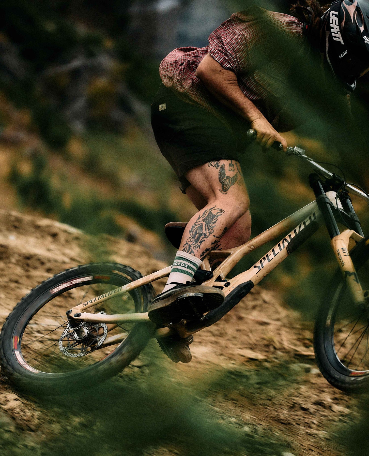 A mountain biker riding a full-suspension bike on a dirt trail, with the rider's legs and tattoos visible and the bike frame showing the 'Specialized' brand name.